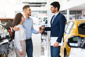 Obraz premium Cheerful man sales associate giving happy young middle eastern couple key from brand new car and smiling, side view. beautiful arabic family buying brand new auto at luxury dealership salon