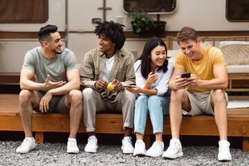 Happy diverse friends sitting near motorhome, talking to each other, browsing web on smartphone, spending time together on camping trip. Cool young people having fun summer vacation