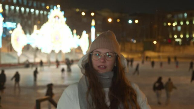 Young woman dressed in warm winter clothes waves affably at camera. Girl was skating at ice rink in city park at night, surrounded by Christmas lights.