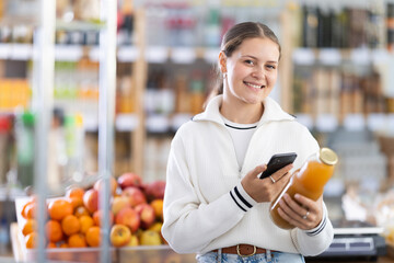Young woman of European appearance scans the label on a bottle of peach juice. Supermarket visitor buying fruit juice with qr code