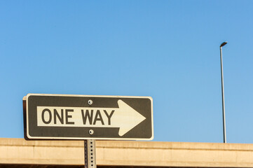 One-way traffic sign with a white arrow pointing right, positioned near a freeway overpass and...