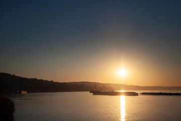 Cargo ships traverse the Danube at dusk, where the evening light casts a serene glow over the water and industrial backdrop of this riverside city.