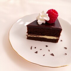 A slice of chocolate cake with a layer of cream in the center, decorated with whipped cream and raspberries on top, on a white plate with a gold rim.
