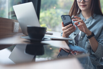 Young asian business woman using mobile phone and working on laptop computer at coffee shop. Woman freelancer online working, surfing the internet at cafe, business casual