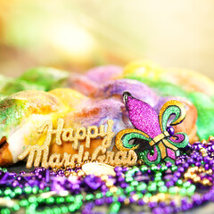 Happy Mardi Gras text in gold glitter and a king cake with yellow, green, and purple sprinkles surrounded by Mardi Gras beads and a glittering fleur de lis. © Leigh Prather