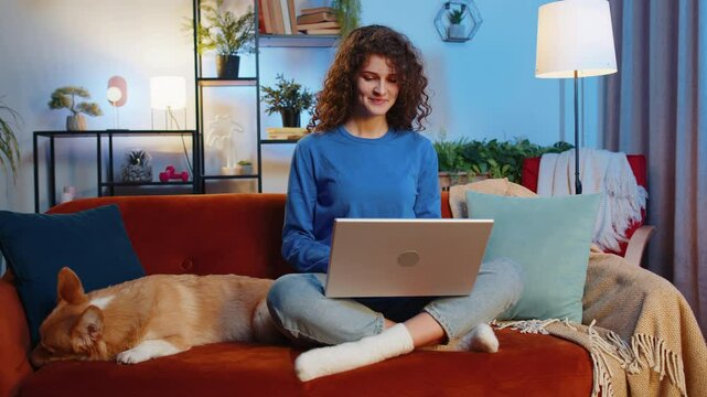 Beautiful young female girl freelancer with curly hair in casual clothes using laptop sitting beside corgi dog on sofa in living room. Positive woman with netbook working on online project at home.
