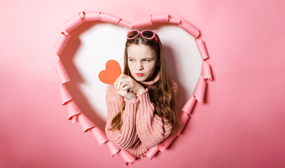 Cheerful young girl poses against pink heart-shaped background, holding heart card celebrate Valentines Day or Womens Day. Smile radiates love, friendship and joy, perfect for 14 February festivities