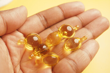 Hand holding several golden capsules with clear liquid against a bright yellow background