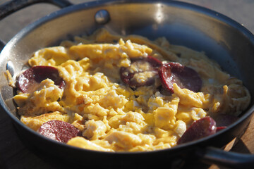 Scrambled eggs with sausage cooked outdoors in a skillet during sunny weather