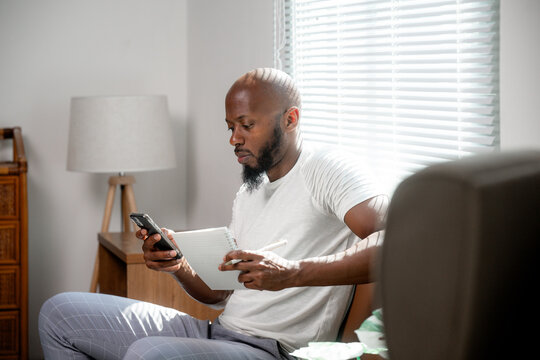 Young african american man working from home, using smartphone and taking notes on notepad, sitting on sofa in living room - Powered by Adobe