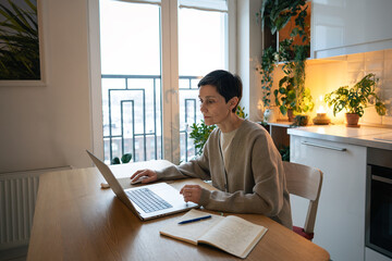 Weary concentrated mature woman taking notes in notebook while remote working on laptop at home. Busy serious woman online learning, do remote tasks, overloaded schedule and organizing timetable