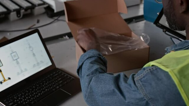 Over-shoulder shot of African American male quality controller inspecting product sample from new batch at factory, taking industrial LED lamp from box and comparing to picture on laptop