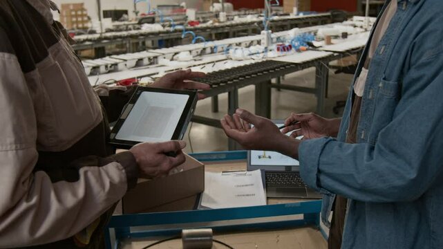 Medium rear shot of two unrecognizable multiethnic male engineers at industrial lighting factory standing by laptop with technical drawing, discussing specifications for new lamp to fulfill order