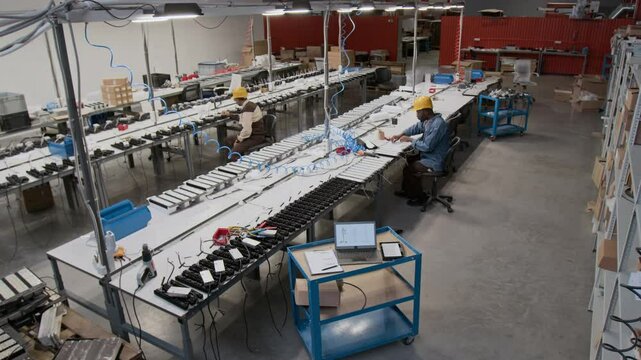 Wide high-angle security camera shot of multiethnic factory specialists working in spacious workshop, assembling industrial lamps, African American man walking over to laptop to check specifications