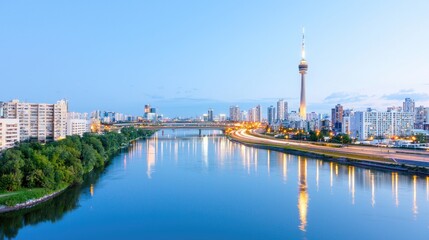Fototapeta premium Modern city skyline with a tall television tower reflecting in the calm river during blue hour.