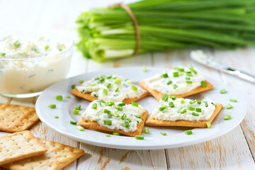 Crispy crackers and a glass bowl with a cream cheese spread and cut chives on a light kitchen table.