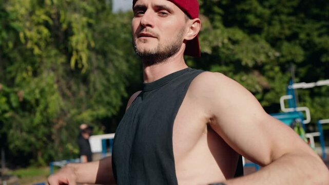 A muscular man is flexing his biceps on an outdoor sports field. He wears a black tank top and a red baseball cap. He shows off his muscles after a workout.