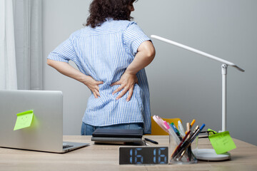 A woman in a striped shirt stands at a cluttered desk, holding her lower back in discomfort. The desk has a laptop, clock, lamp, and office supplies. The scene highlights back pain and work fatigue.