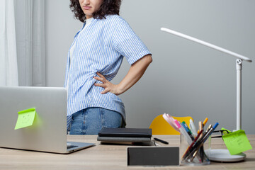 A woman in a striped shirt stands at a cluttered desk, holding her lower back in discomfort. The desk has a laptop, clock, lamp, and office supplies. The scene highlights back pain and work fatigue.