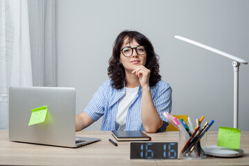 A woman sits at a desk, appearing thoughtful and slightly bored, resting her head on her hand. Wearing glasses and a striped shirt, she is surrounded by a laptop, stationery,