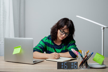 A woman in glasses and a green-striped shirt sits thoughtfully at a desk, her chin resting on her hand. A laptop, pens, and a digital clock showing 16:16 are arranged neatly on the desk