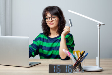 A woman wearing glasses and a green-striped shirt sits at a desk with a laptop. She holds a credit card in her hand and smiles while looking at the screen. The workspace is neat and modern