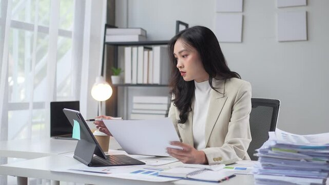 A woman is sitting at a desk with a stack of papers in front of her. She is reading a piece of paper and she is focused on the task at hand. Concept of productivity and concentration