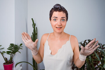 A surprised woman with dark hair covered in dye, wearing gloves and a light camisole, stands in a brightly lit room with potted plants. She raises her hands, showing messy gloves, and looks amused