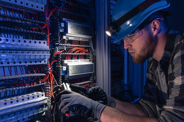 A male electrician works in a switchboard in overalls against the backdrop of emergency lighting.