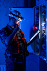 Electrician in overalls, focused on work in switchboard with fuses, using tablet.