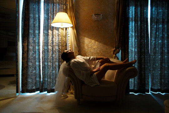 woman sitting on a chair in a room with dim lighting. She is dressed in a white robe and veil, her head is thrown back and her legs are stretched out on the armrest of the chair. - Powered by Adobe