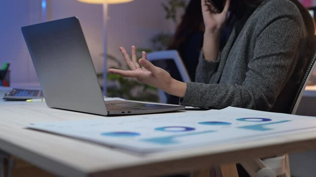 A woman is talking on her cell phone while sitting at a desk with a laptop and a stack of papers