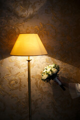 The lamp stands against the background of a wall with patterned wallpaper in golden tones. On the right side of the frame you can see a hand holding a bouquet of white roses.