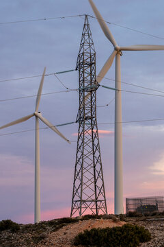 A striking image of wind turbines along with a power transmission tower silhouetted against a colorful sunset sky, showcasing renewable energy infrastructure in Zaragoza Spain