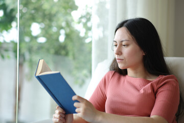 Serious asian woman reading a paper book on a sofa at home