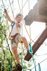 A child in a rope park in nature learns to rock climb. A boy in a helmet walks along a ropeway overcoming obstacles.