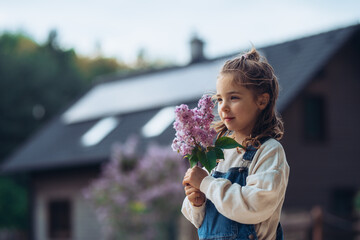 Beautiful young girl with purple lilacs flower in hands. House with solar panels in background.