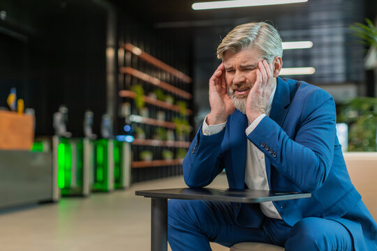 Tired ill middle-aged businessman suffering from headache problem tension and migraine, stress in modern office lobby. Handsome freelancer businessperson man guy sitting in hotel hall. Overworking.