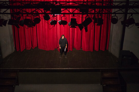 Actor with script standing on theatre stage in front of red curtain