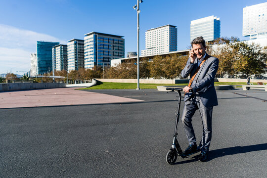 Mature businessman commuting in the city with his kick scooter, usine smartphone