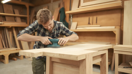Young carpenter sanding wood piece in workshop in furniture factory