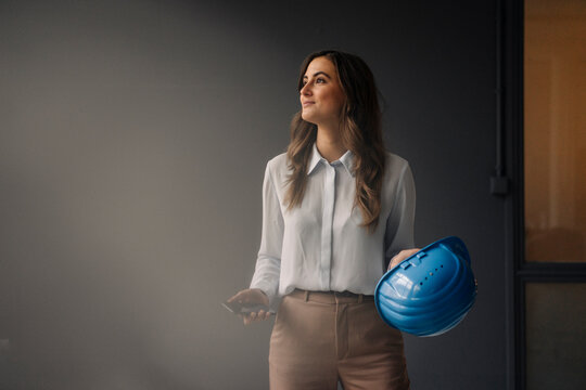 Smiling young businesswoman holding hard hat and cell phone