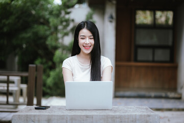 Cheerful Asian woman working in a coffee shop using her laptop. She enjoys teleworking, studying or surfing the internet to relax.