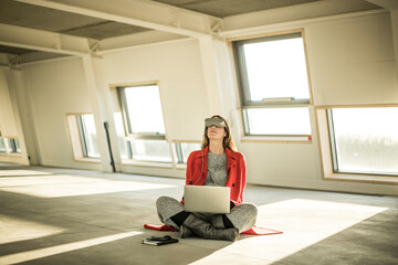 Pregnant busnesswoman sitting on floor of new office rooms, using VR goggles and laptop