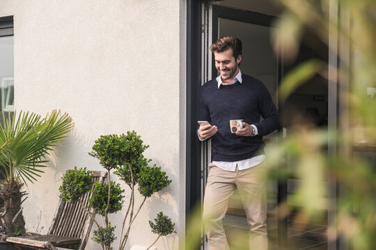Young man leaning in door of his house, holding cup of coffee, using smartphone - Powered by Adobe