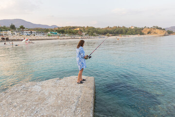 Woman fishing on a pier, gazing over a serene beach resort, calm waters, and distant hills.