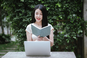 Fototapeta premium A confident young Asian woman is working on her laptop in a vibrant park. E-banking and career aspirations in a modern lifestyle.