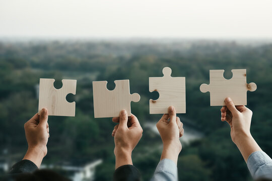 Concept of business people working as a team, cooperation within an organization, hands holding a jigsaw puzzle above their heads with a background of mountains with sunlight shining down.