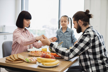 Caucasian family of three holds hands praying before breakfast. Includes father, mother, and young daughter. Table includes healthy food options, evoking warmth, connection, and gratitude.