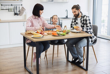 Caucasian family with parents and young daughter enjoying breakfast with pizza, fruit, and salad at home kitchen table. Warm family bonding and morning routine.